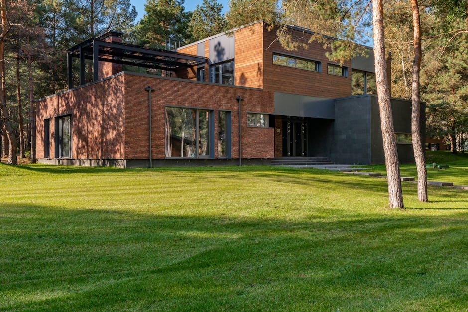 A modern two-story house with a combination of red brick and wooden cladding exterior, set amidst a well-maintained lawn and surrounded by tall, mature trees. The front yard features lush, green grass